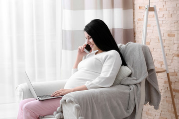 Attractive young pregnant woman with laptop speaking by cellphone at home