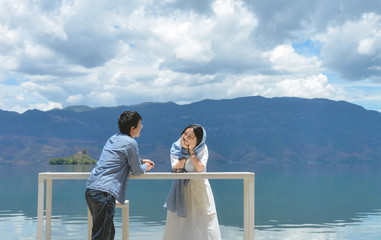 Happy young Chinese couple leans on white long table looking at each other, by the bank of Lugu...