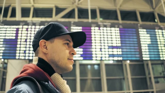 Caucasian man wearing cap walks near airport departure board. Tourism or travel concepts. 4K steadicam video