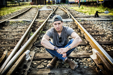 Fototapeta premium Attractive young man sitting on railroad, wearing grey t-shirt, baseball cap and jeans, looking at camera