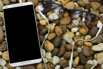 White smartphone with blank screen and earphone and eyeglasses on stones background.