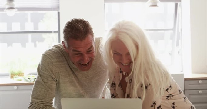 Excited senior couple looking at a laptop together in the kitchen pointing and smiling at something amusing on the screen