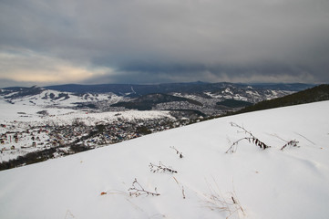 Winter mountain hill slope field with snow on the background of village town in dark cloud gloom...