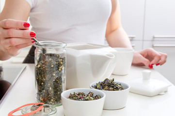 Young woman prepares brew green tea on white table with different kinds of herbs