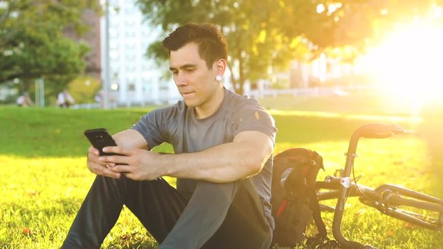 Man with bike relaxing at park in Chicago. Young caucasian man sitting on the grass and typing on the phone. Backlight view with traffic blurred on background