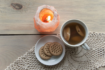 Cozy home still life. Candle of pink salt with fire, a cup of apple compote and plate with cookies on knitted napkin on a light wooden background. Top view.