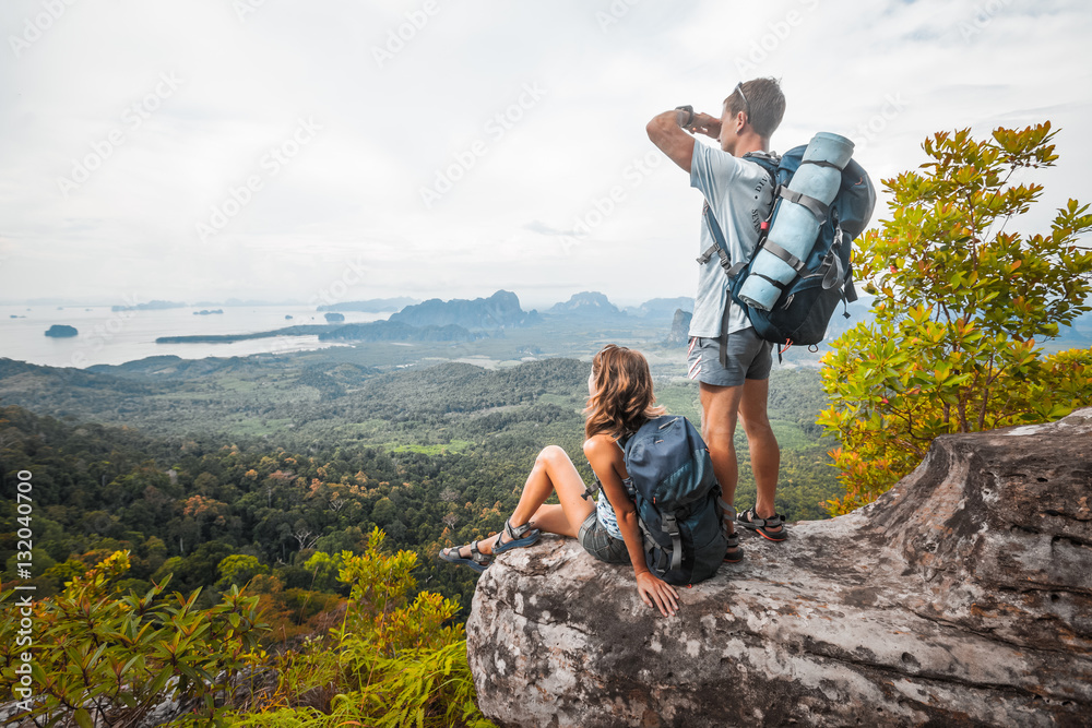 Poster couple of tourist with backpacks relaxing on top of a mountain and enjoying the view of valley