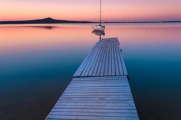 Sunset over lake with yacht, moored by wooden pier.