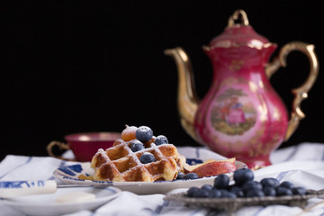 Blueberry apple waffle with honey set on a dish decorated with fresh blueberry, apple, tea pot set ready to serve.