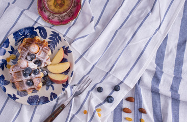 Blueberry apple waffle with honey set on a dish decorated with fresh blueberry, apple, tea pot set ready to serve.