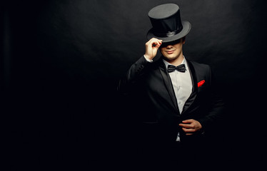 A magician in a black suit holding an empty top hat and magic wand isolated on black background - obrazy, fototapety, plakaty