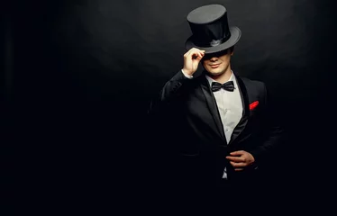 Fotobehang A magician in a black suit holding an empty top hat and magic wand isolated on black background © Cheberkus