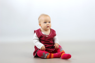 Naklejka premium Children, Family concept. Cute little baby girl in red dress sitting on the floor and looking sideways. 