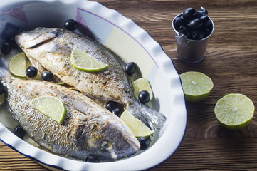Fresh uncooked dorado with lemon, lime, olives, oil and spices on rustic wooden board. Raw fresh fish with seasonings in roasting pan before baking on brown wooden background.
