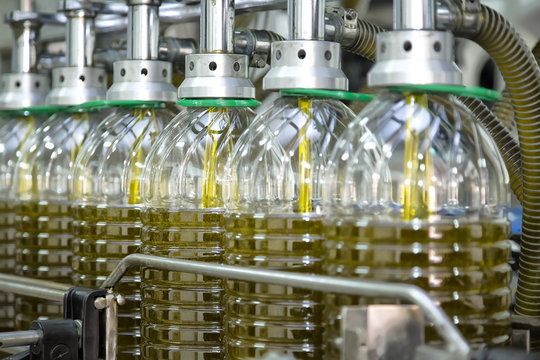 Envasado de garrafas de aceite de oliva virgen producci&oacute;n industrial en almazara moderna llen&aacute;ndose autom&aacute;ticamente en proceso alimentario con maquinaria automatizada de industria agroalimentaria