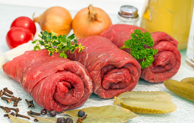 Drei rohe Rinderrouladen aufgerollt, umgeben von Tomaten,Gurken, Zwiebeln und Gewürzen