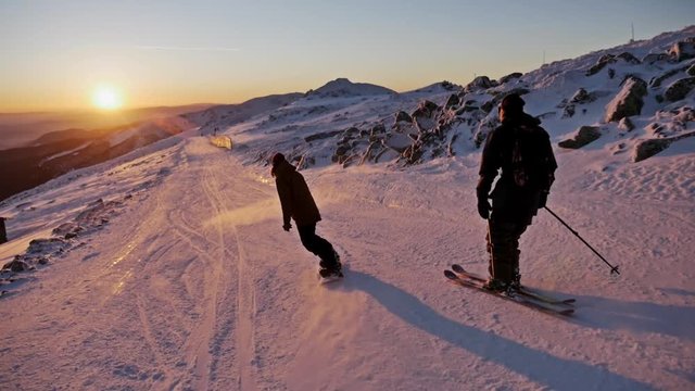 Slowmotion footage of a skier and snowboarder riding the slope during a beautiful sunset.