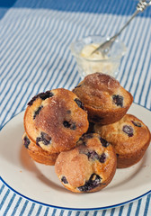 Blueberry muffins with cinnamon sugar on a round white plate with a blue rim on a blue and white striped towel with a clear glass cup of butter and a silver spreader