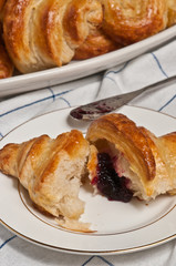 Fresh, baked croissant with raspberry jam spread on inside with a silver spreader on a round white dish with a gold rim sitting on a white table cloth with blue stripes