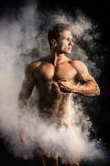 Fototapeta na wymiar Totally naked male bodybuilder with smoke hiding genitalia, looking away to a side, on dark background