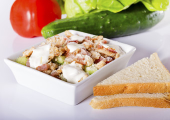 Simple, fresh salad with vegetables, grilled chicken and smoked ham in white bowl next to toast, cucumber and tomato.