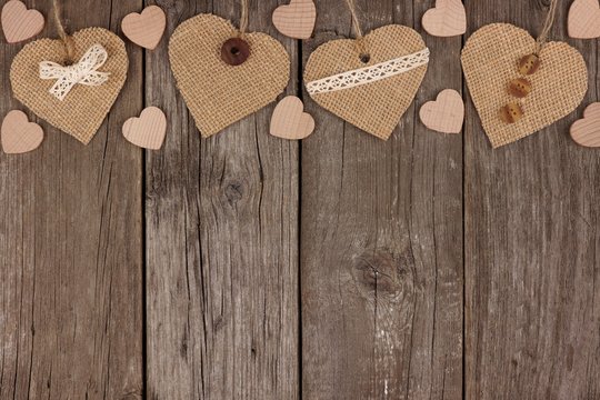 Top border of handmade burlap hearts with ribbon and buttons over a rustic wooden background