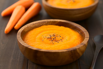 Fresh homemade cream of carrot soup with freshly ground black pepper on top served in wooden bowls, photographed on dark wood with natural light (Selective Focus, Focus in the middle of the soup)