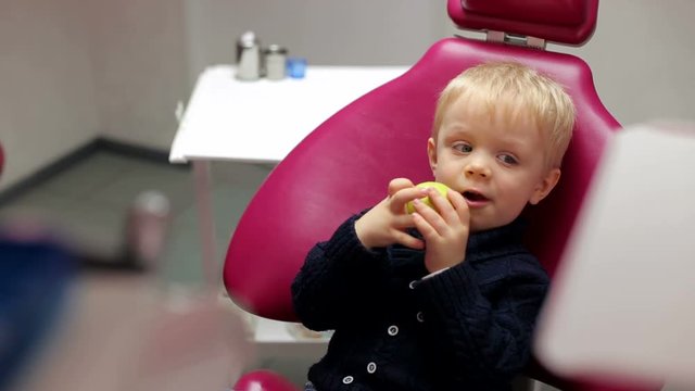 Small child sitting in the dental chair and eating a green apple. 