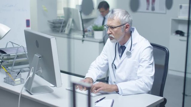 Senior Doctor and His Assistant Working at Desktop Computers. Sitting in Brightly Lit Office.  Shot on RED Cinema Camera in 4K (UHD).