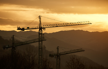 Three construction cranes in front of mountains during sunset.