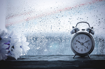Flower and retro alarm clock on table in front the rain - 132107365