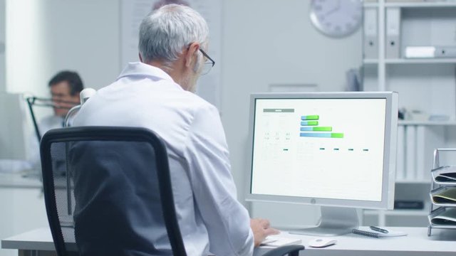 Senior Male Laboratory Researcher Working with Graphs on His Personal Computer. His Assistant Works at His Desk in the Background.  Shot on RED Cinema Camera in 4K (UHD). 