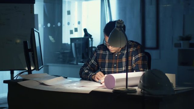 Young Capable Architectural Engineer Working Late Hours in His Office. Office is Dark Only His Table Light is On. Various Plans and Blueprints Lying on the Table. He Draws.