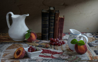 old books , white jugs , grapes , and peach on a colored carpet