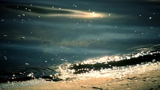 Surf with blue water glittering in sun at dawn or sunset with sparkles from sunshine and small waves in sea, river, lake or ocean with soft light