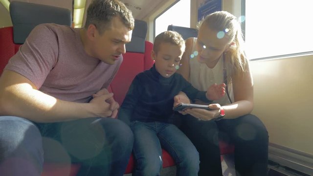 View of happy young family, mother, son and father, riding in the train against window, sit on their seat places and watching smartphone and speaking. Prague, Czech Republic