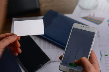 Businessman in the office standing near the table with the papers with charts, tables, mobile phone and laptop and holding a business card (visit card). Blank, free space