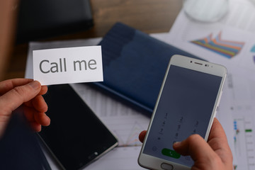 Businessman in the office standing near the table with the papers with charts, tables, mobile phone and laptop and holding a business card (visit card) with text call me
