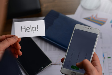 Businessman in the office standing near the table with the papers with charts, tables, mobile phone and laptop and holding a business card (visit card) with text help