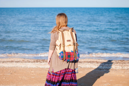 A girl stands back and looks at the sea. Hippie Girl looks at the sea. Woman with a bottle of wine is by the sea. hippie girl with an old backpack and a bottle of wine.