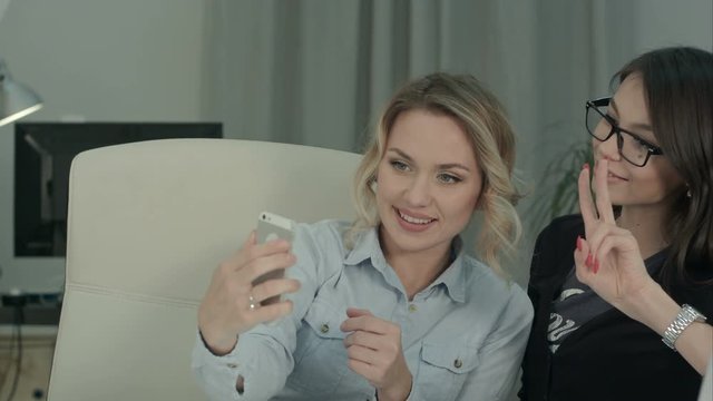 Two female colleagues taking selfies with phone sitting at the desk