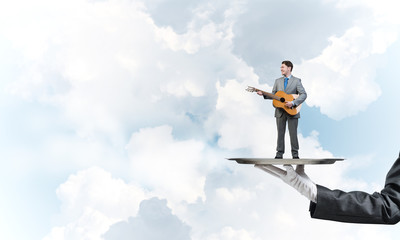 Businessman on metal tray playing acoustic guitar against blue sky background