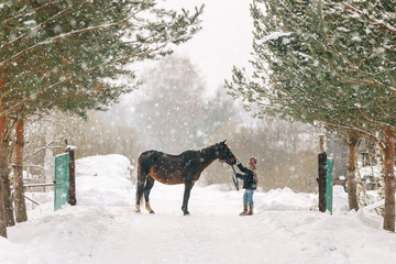 Young girl child in a fur coat and scarf in winter standing on the snow in front of the gate of a horse and stroking its nose