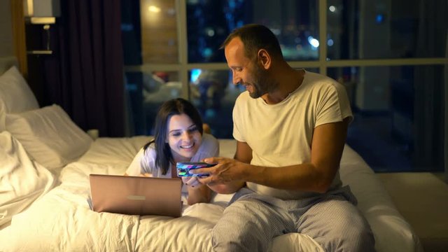 Young, happy couple with smartphone and laptop talking in bedroom at night
