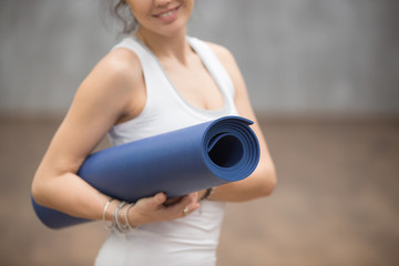 Attractive woman wearing white sportswear holding blue yoga or fitness mat after working out at home or in club. Friendly smiling sport instructor with yogamat. Hands close up photo