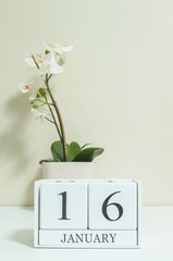 Closeup white wooden calendar with black 16 january word with white orchid flower on white wood desk and cream color wallpaper in room textured background , selective focus at the calendar