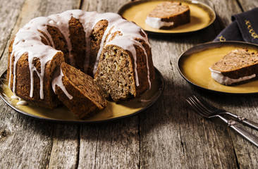 Banana cake with apples and walnuts drizzled frosting and sliced pieces on plates with effect craquelure on the shabby wooden background. Selective focus 