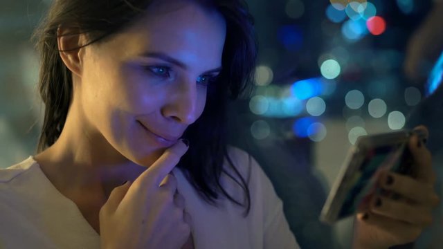 Happy, young woman watching movie on smartphone by window at night
