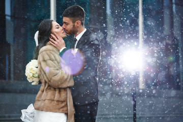 Beautiful and gentle winter wedding photo session outdoors of the elegant couple (bride in a white dress with veil holding a bouquet and groom in the classic tuxedo costume), family romantic portrait