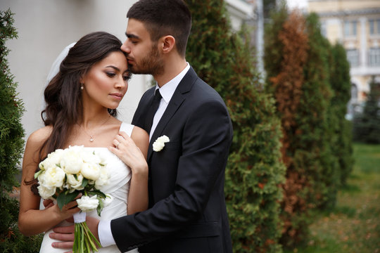 Beautiful and gentle wedding photo session outdoors of the elegant couple (bride in a white dress with veil holding a bouquet and groom in the classic tuxedo costume), family romantic portrait